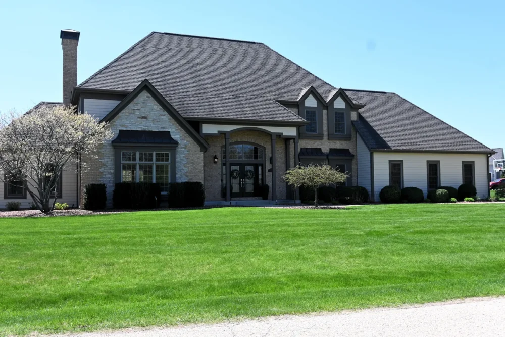 Exterior painting project on Waterleaf Ln in Hartland WI featuring a large home with siding, dark trim, dormers, stone accents, and a green front lawn.