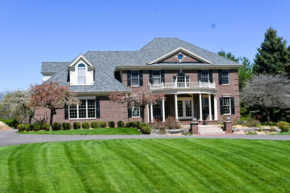 Exterior painting project on Woodland Hill Dr in Delafield WI featuring a large brick home with black shutters, white columns, balcony details, and a green front lawn.