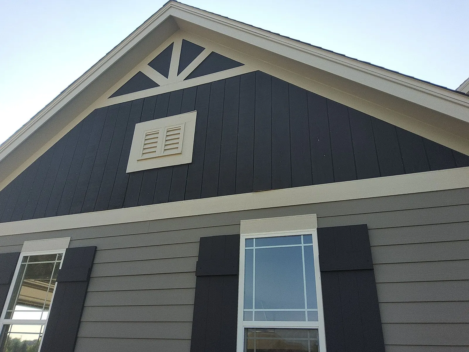 Dark board-and-batten gable with cream trim above gray siding and shuttered windows.